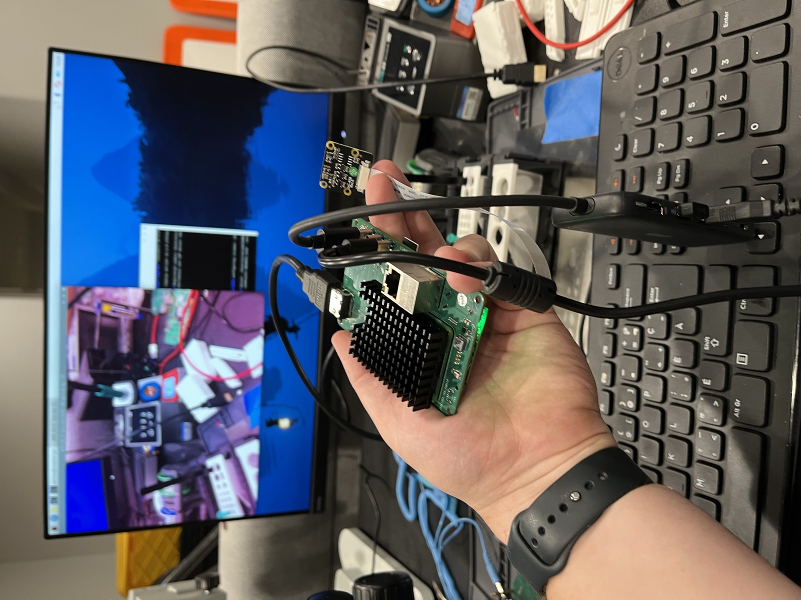 The assembled board held in one hand — a heatsinked CM5 on top, HDMI and USB cables in the back edge, a camera module on a ribbon into one of the MIPI connectors, and the green power LED lit. Behind it, the monitor the board is driving, with the camera's live preview open on it.